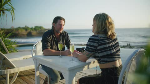 A man and a woman are sitting at a table on the boardwalk having a drink as the waves roll in