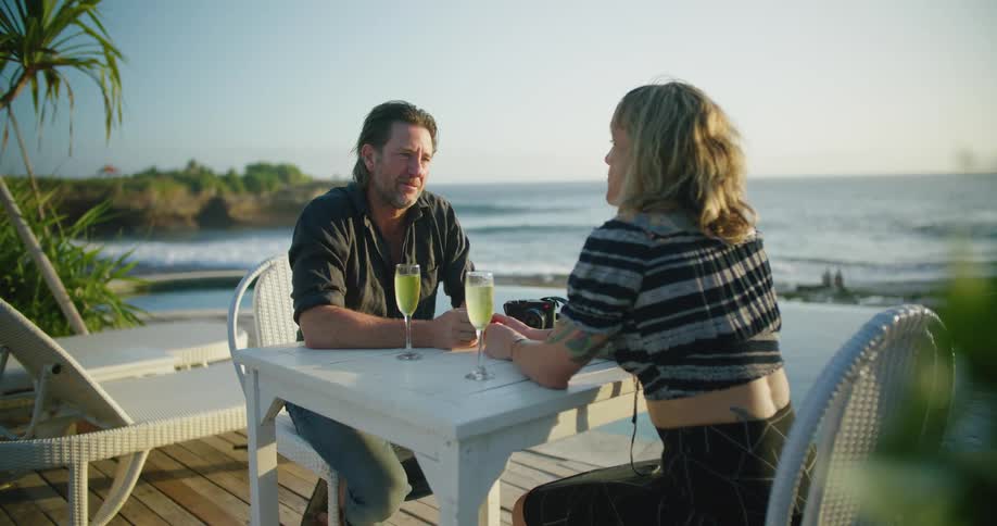 A man and a woman are sitting at a table on the boardwalk having a drink as the waves roll in