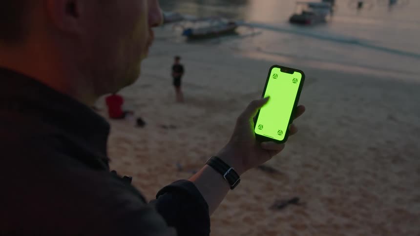 Man stares at a smartphone with a green replaceable screen he is holding on his hand on a beach at sunset