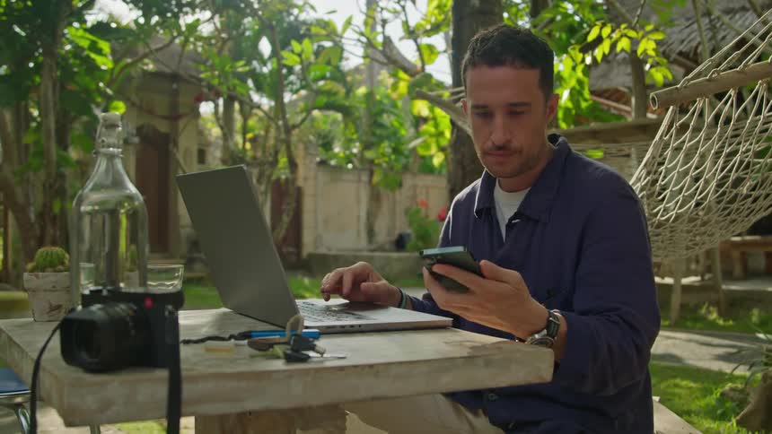 Photographer sitting on an outdoors tropical restaurant working on a laptop while checking the phone