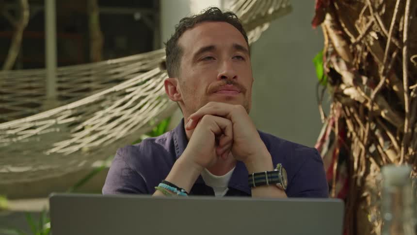 A man is sitting at a table outside typing on a laptop computer and he stops to think resting his chin on his hands