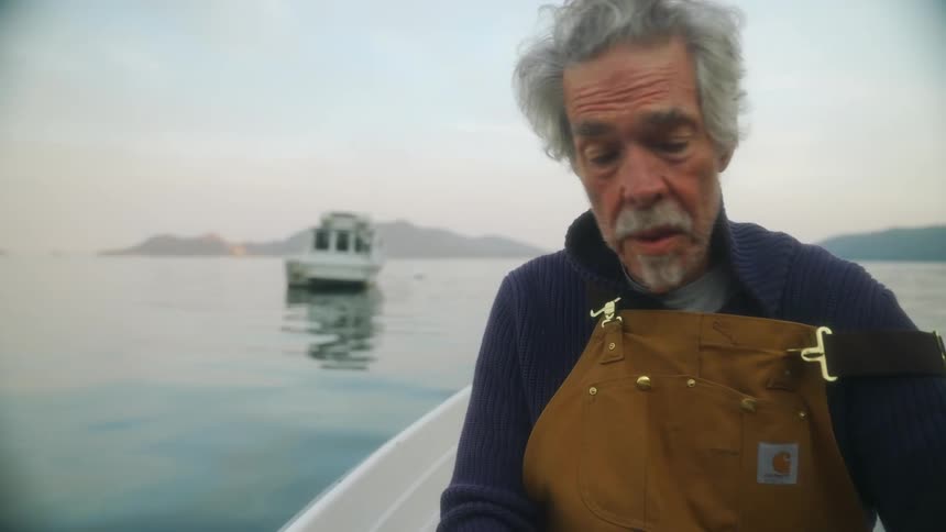Elderly fisherman wearing an apron sits on a boat on the calm surface of a lake at morning