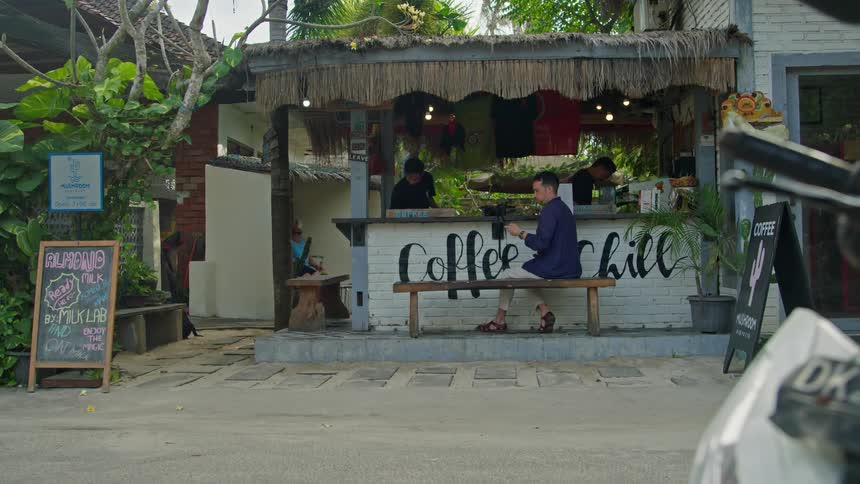 Two baristas working in an open coffee bar while a customer sits at a bench near the road