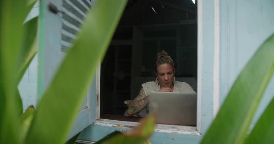 Young tattooed woman sitting near a window on a tropical house works on a laptop