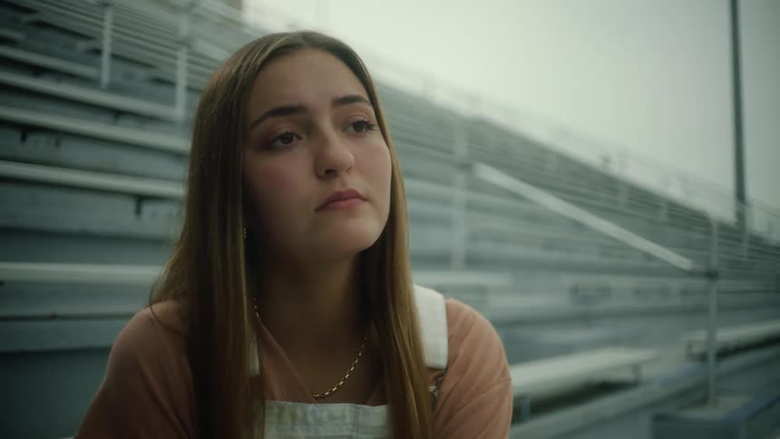 A teenage girl is standing alone near empty bleachers as her hair moves in the breeze