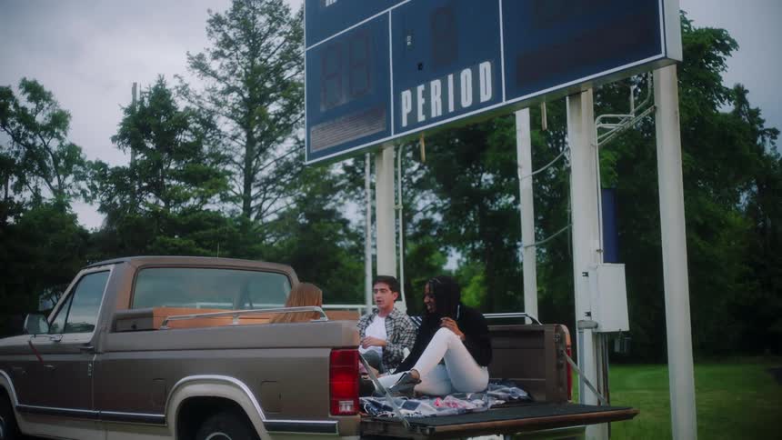 Three friends sitting in the bed of a pickup parked under the scoreboard
