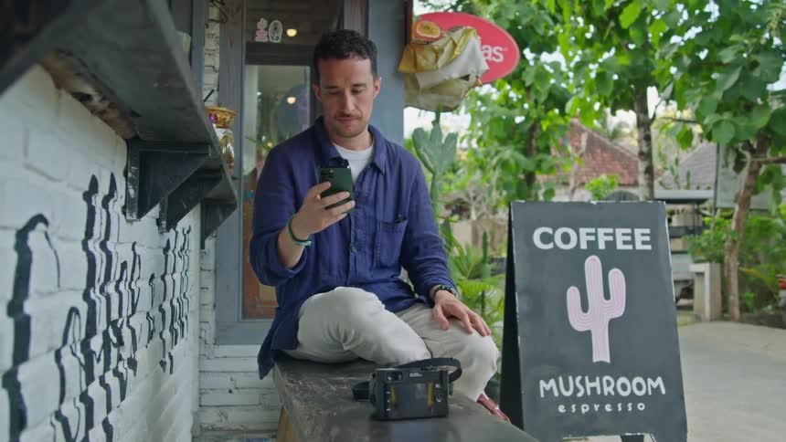 A man is sitting on a bench at an outdoor cafe using his cell phone as a motorbike passes by on the street