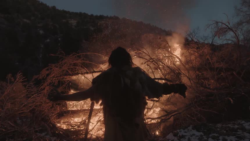 A man standing in front of a large fire with branches burning outside out at night