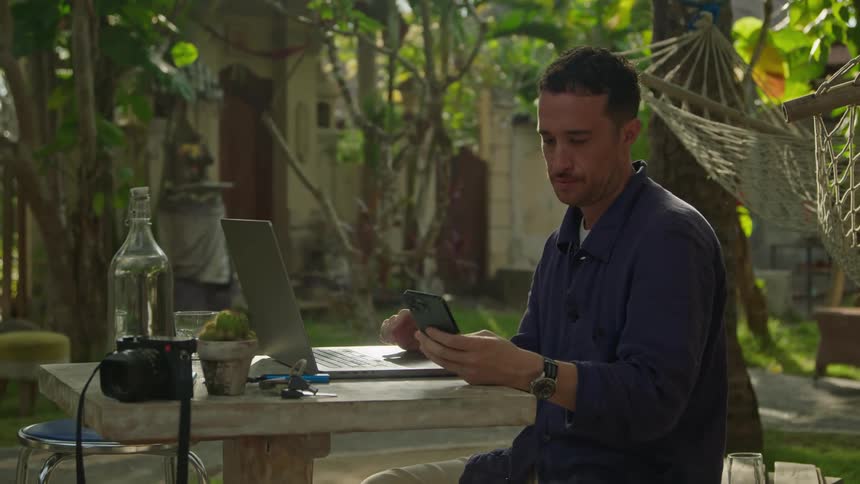 A man is sitting at a table outside typing on a laptop computer and he stops to check his phone and then he goes back to typing