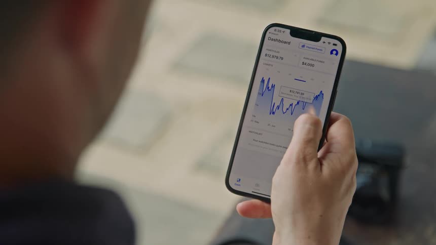 A man is holding his cell phone and using his thumb to move across a graph on the screen