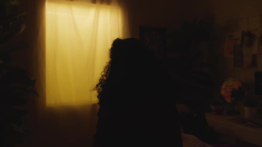 Young teenage woman sitting on a dimly lit room looking at the curtains on the window