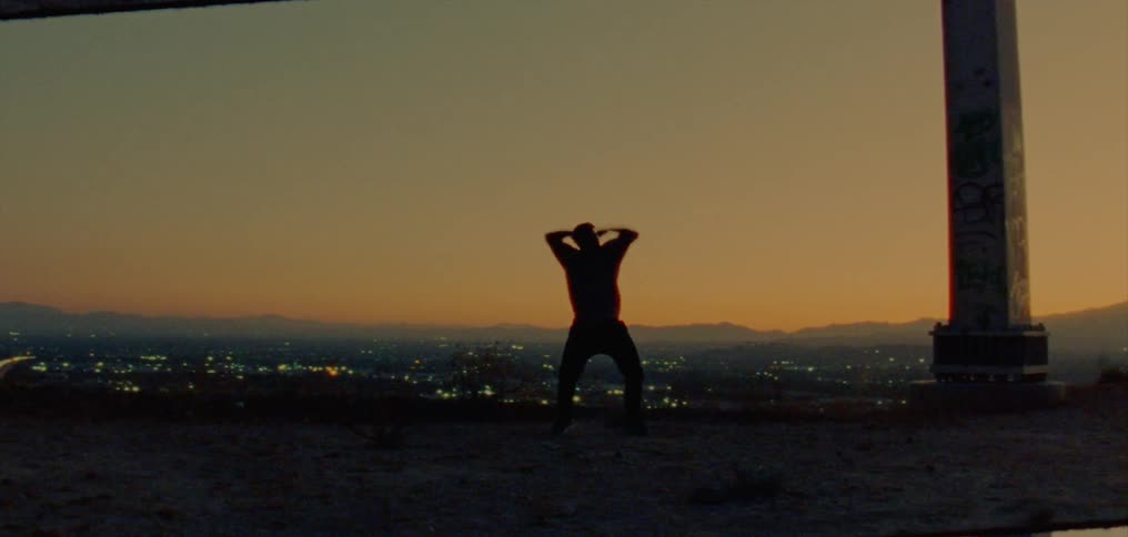Silhouetted against a red sky male dancer dances near structure support column on hill overlooking city