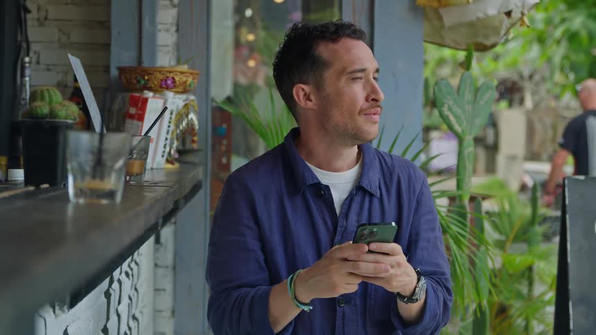 A man looks up from his phone to smile and wave while sitting at an outdoor bar