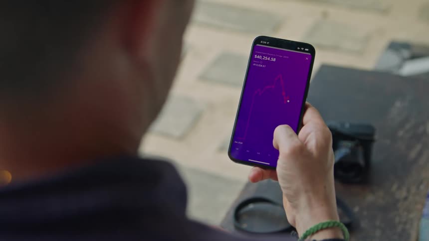 A man is holding his cell phone and using his thumb to move across a graph on the screen