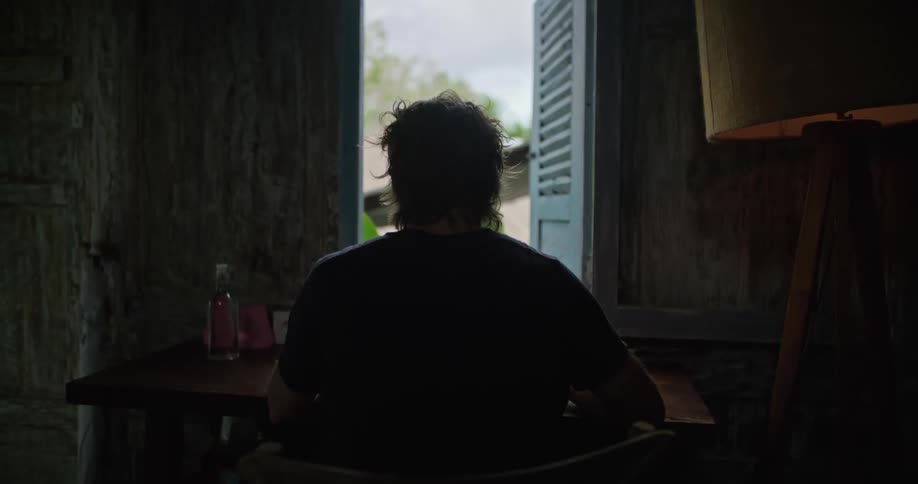A disheveled looking man sits at a table with his back to the camera doing something with his hands in a dingy looking room