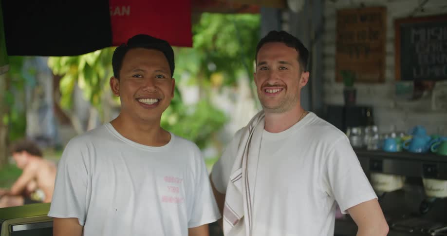 Two men behind the counter of a tropical bar smile looking at the camera