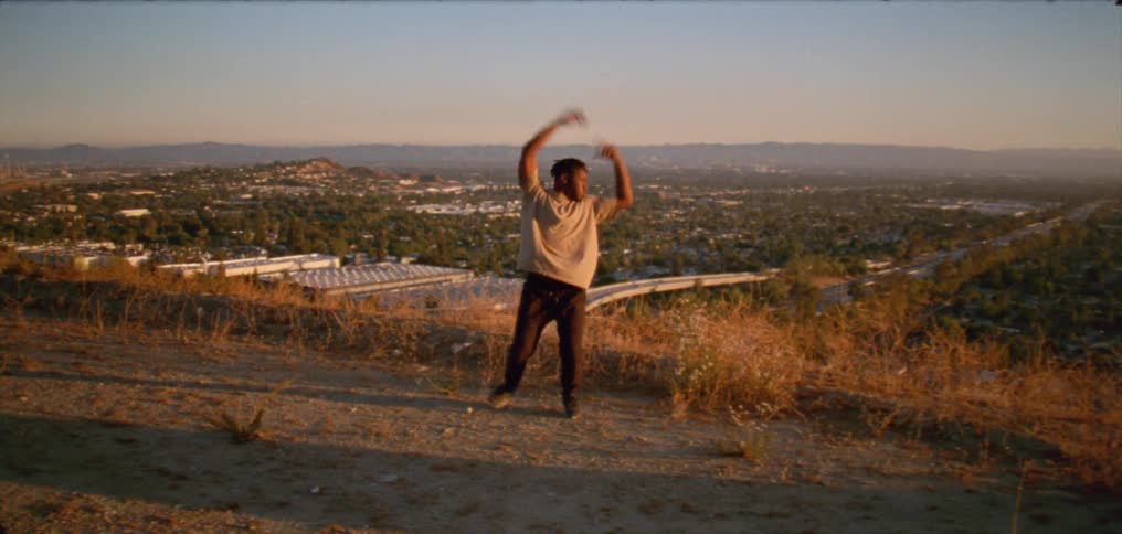 A man is standing on top op a hill dancing as the sun shines upon him and a small town is below with mountains on the horizon