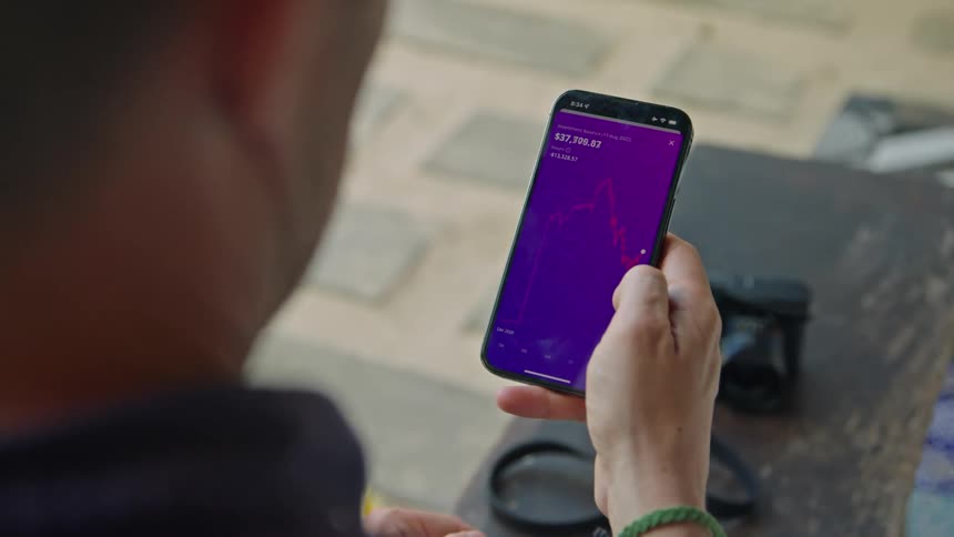 A man is holding his cell phone and using his thumb to move across a graph on the screen