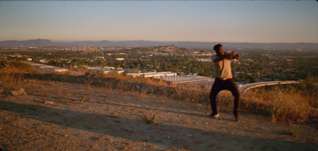 Male dancer with dreadlocks white shirt and black pants dances on hill overlooking city at dusk