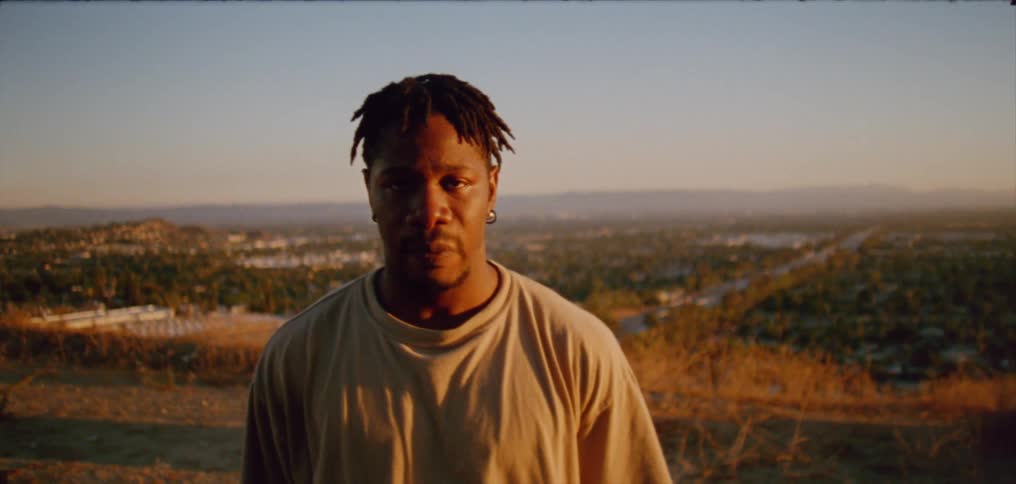 A black man is standing outside looking into the camera while licking his lips