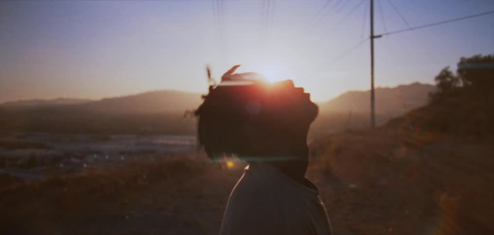 A young man lifts up his head standing in profile with the sun glaring behind him on a hillside trail