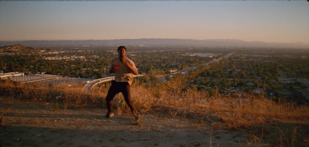 Young man wearing a white shirt dancing on a hill near a small town at evening