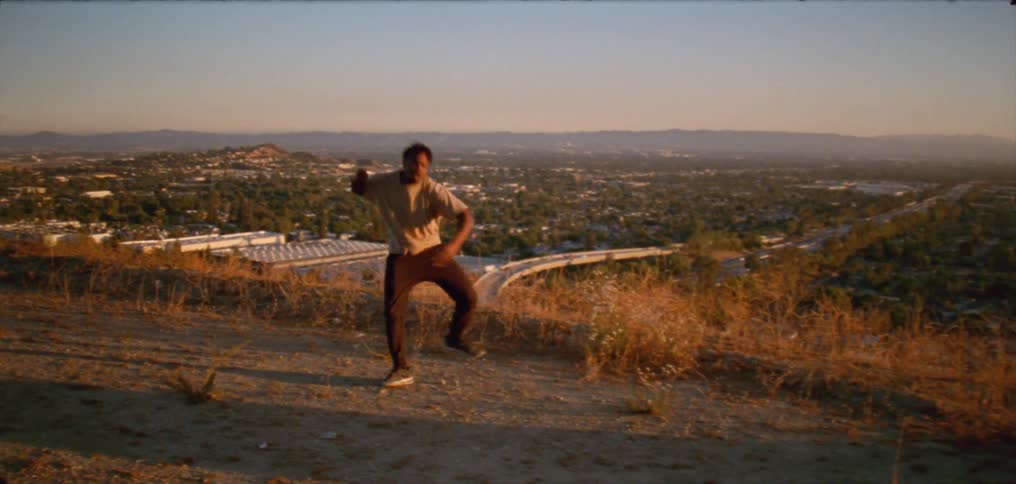 Young man wearing a white shirt dancing on a hill near a small town at evening