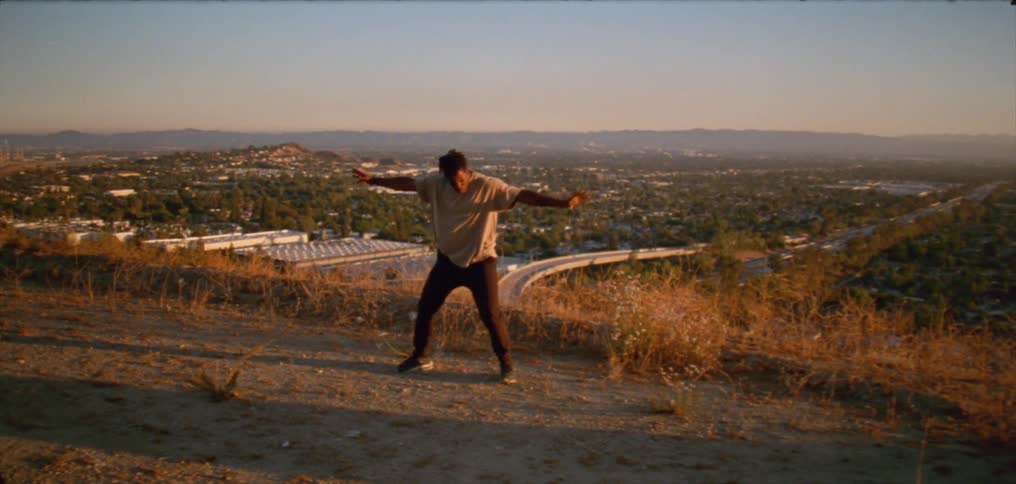 Young man wearing a white shirt dancing on a hill near a small town at evening
