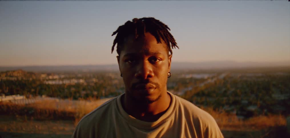 Young man wearing a white shirt stands on top of a hill next to a small town at evening looking at the camera