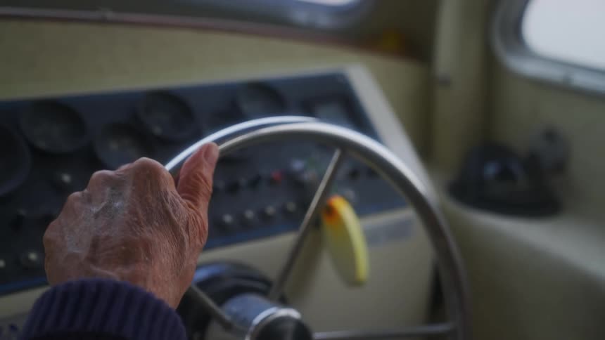 Elderly fisherman turns the wheel of a small fishing boat sailing on a lake