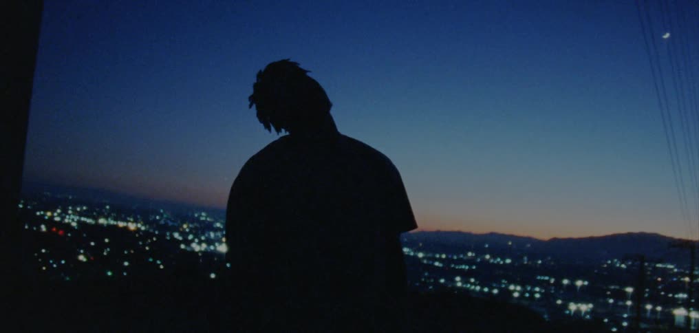 Silhouette of a man standing outside and moving his head as city lights shine behind him and there are mountains in the distance