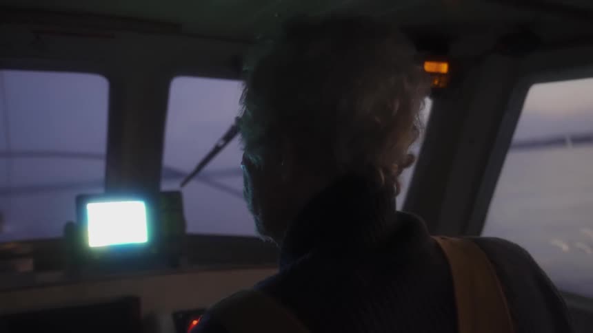 An older man pilots a motorboat across the water under a gray cloudy sky
