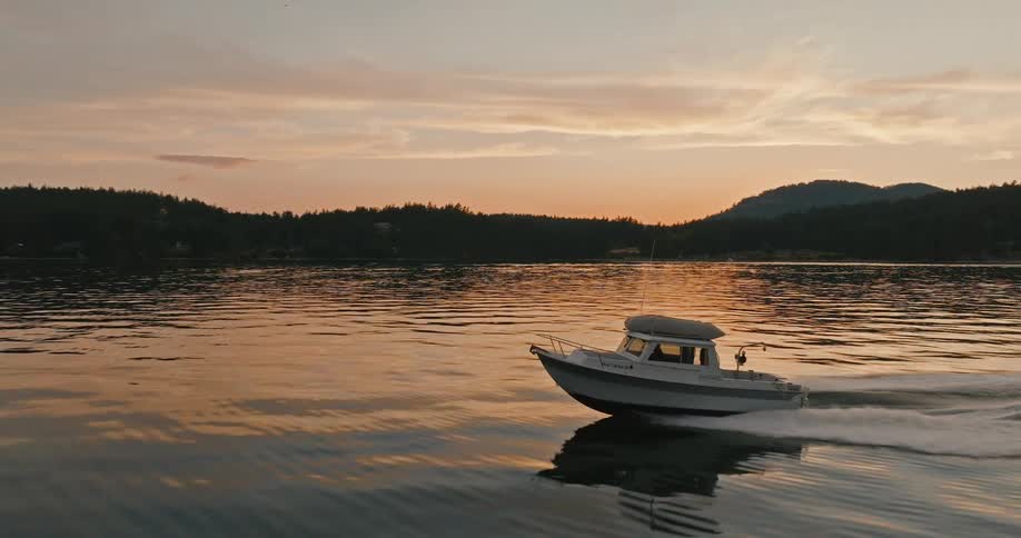 A boat is sailing across the water near shore with a forest along the coast and a mountain in the distance with the sun setting on the horizon