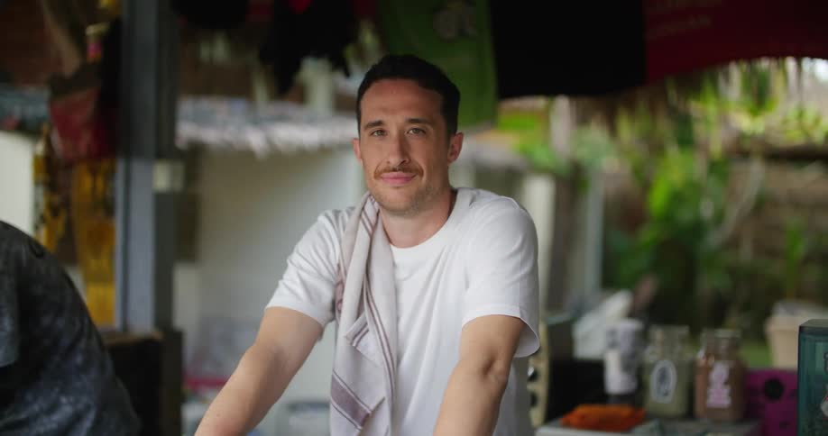 A man is standing at the counter of an outdoor cafe with a smile on his face as he looks around