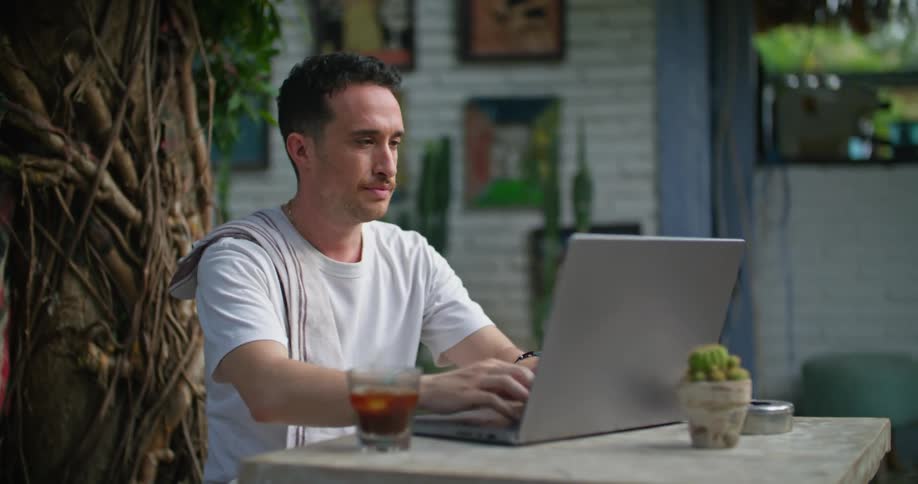 A man is sitting outside at a table with a laptop computer and he stops to reach for his drink