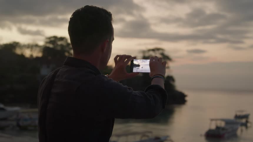 Man standing in front of the coast takes pictures of the ocean with his phone