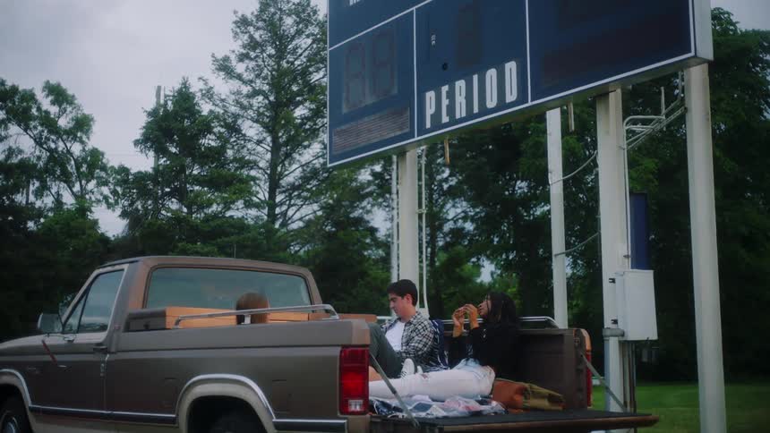 Friends sit in bed of pickup truck parked near sport scoreboard talking and eating