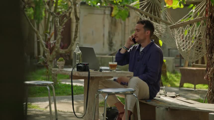 A man is sitting outside at a table sipping his drink and typing on a laptop computer when he answers his phone and looks at his computer as he talks