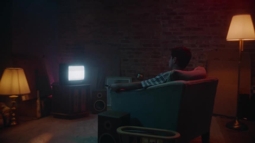 A young man holding a remote sitting in an armchair staring at a small tv playing static