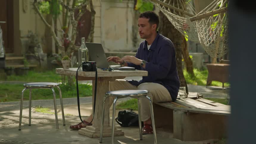 Man sits at outdoor table typing on laptop placed near camera