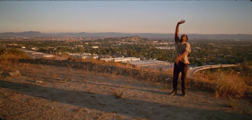 Young man wearing a white shirt dancing on a hill near a small town at evening