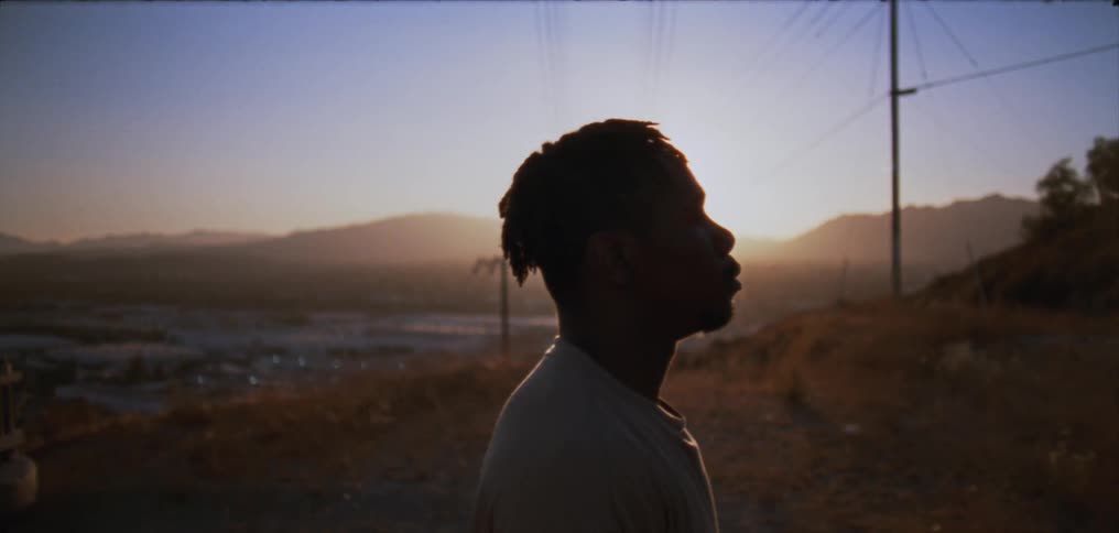 Man stands on hill overlooking town partly silhouetted by sun rising over mountains on horizon