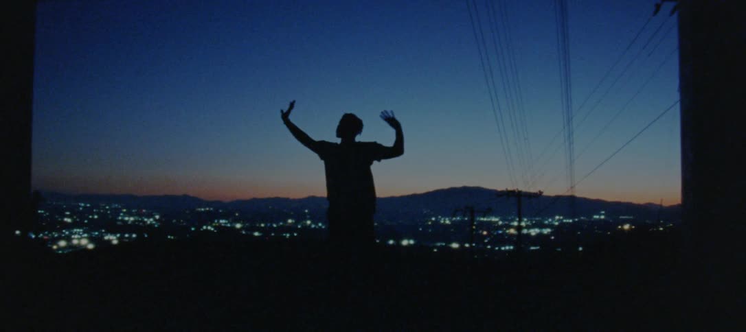 A mans silhouette is shown standing in front of a city skyline at night while waving his arms and hands slowly above his head
