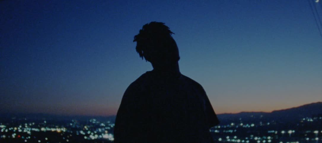 A silhouette of a man is shown standing in front of a city skyline at night while he looks into the camera