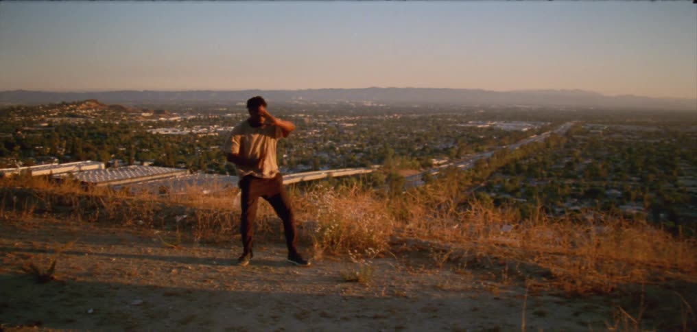 A man dancers atop a hill overlooking a freeway in a rural area in the evening