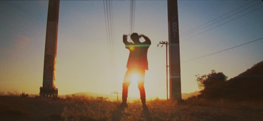 Sun shines behind the silhouette of a man dancing between two poles in the countryside at sunset