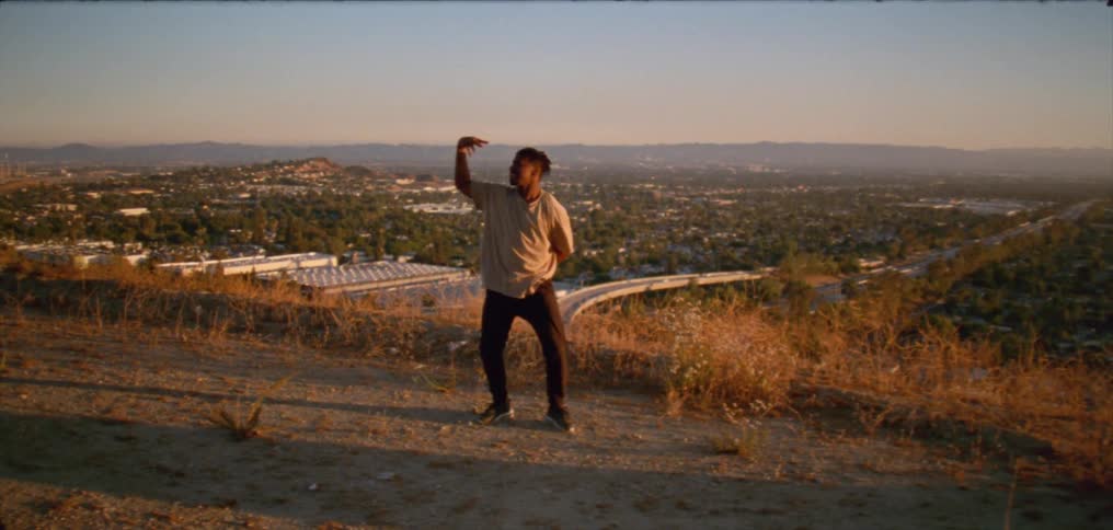 Young man wearing a white shirt dancing on a hill near a small town at evening