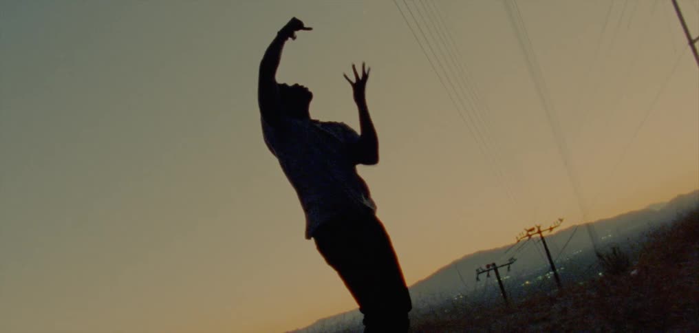 Male dancer partly silhouetted against red tinted sky dances under powerlines with town and hills in the background