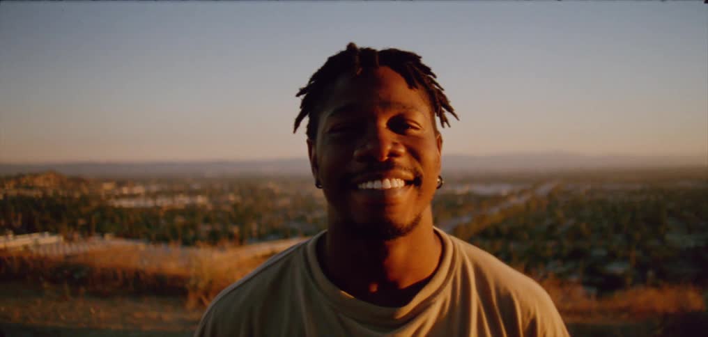 Sunlight shines on the face of a man as he smiles standing outside with neighborhoods below in the distance under a clear sky