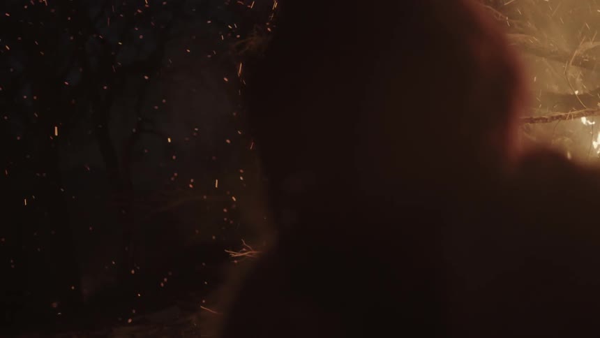 A man standing near a pile of burning branches as embers float into the night sky